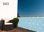 Blauwe balkonafscherming met witte stippen gemonteerd op de terrasreling naast een fauteuil en een plant.