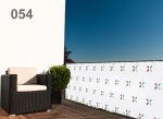 Custom-made raster balcony screen with colourful patterns on a terrace with a woven armchair and green bush.