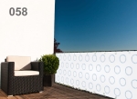 White balcony screen with blue circle pattern on a terrace next to a black armchair and a potted plant.