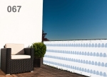 Raster balkonafscherming op maat met blauw-witte patronen, gemonteerd op de terrasleuning naast een fauteuil en plant.