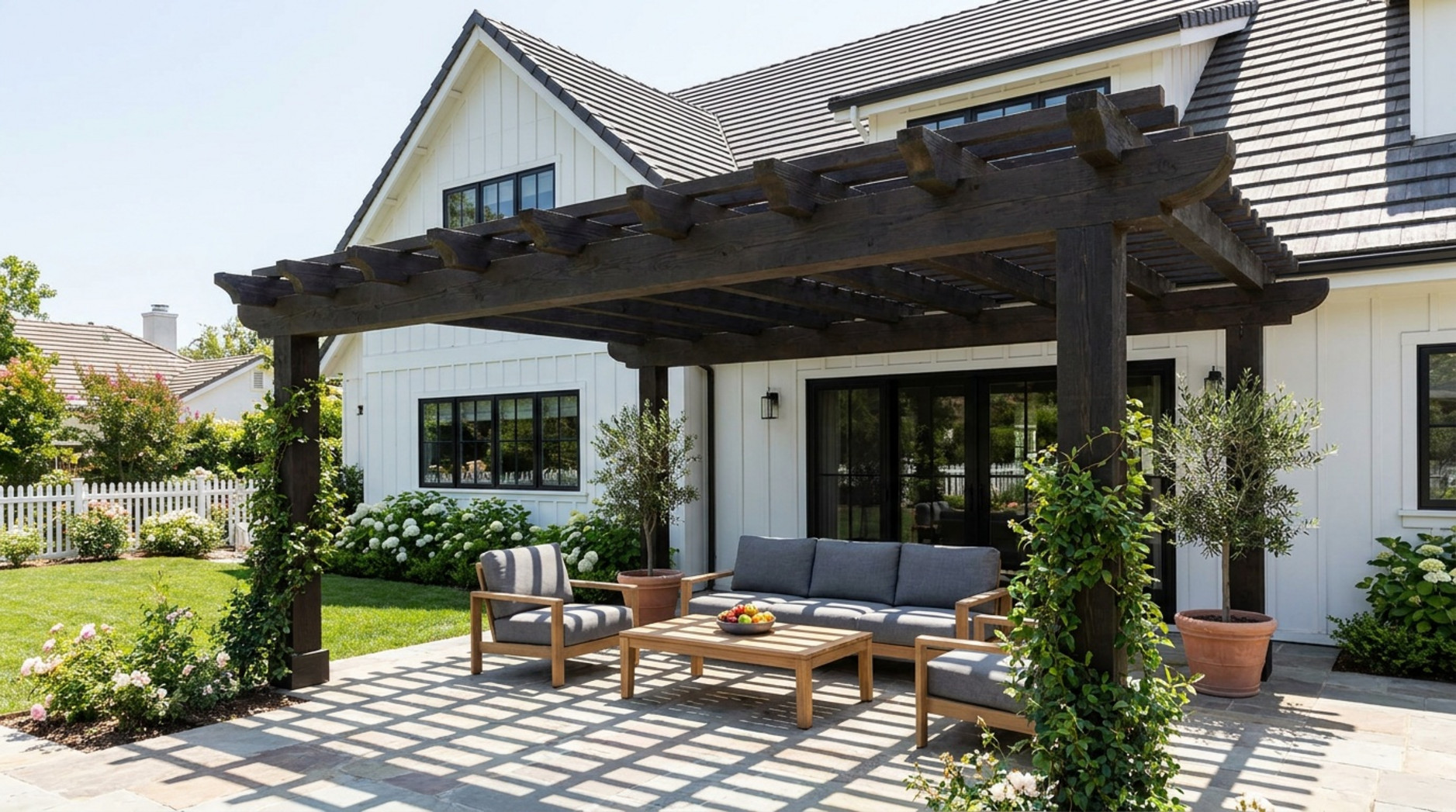 A spacious backyard with a large wooden pergola attached to a modern farmhouse, featuring outdoor seating and sunlight filtering through the rafters.