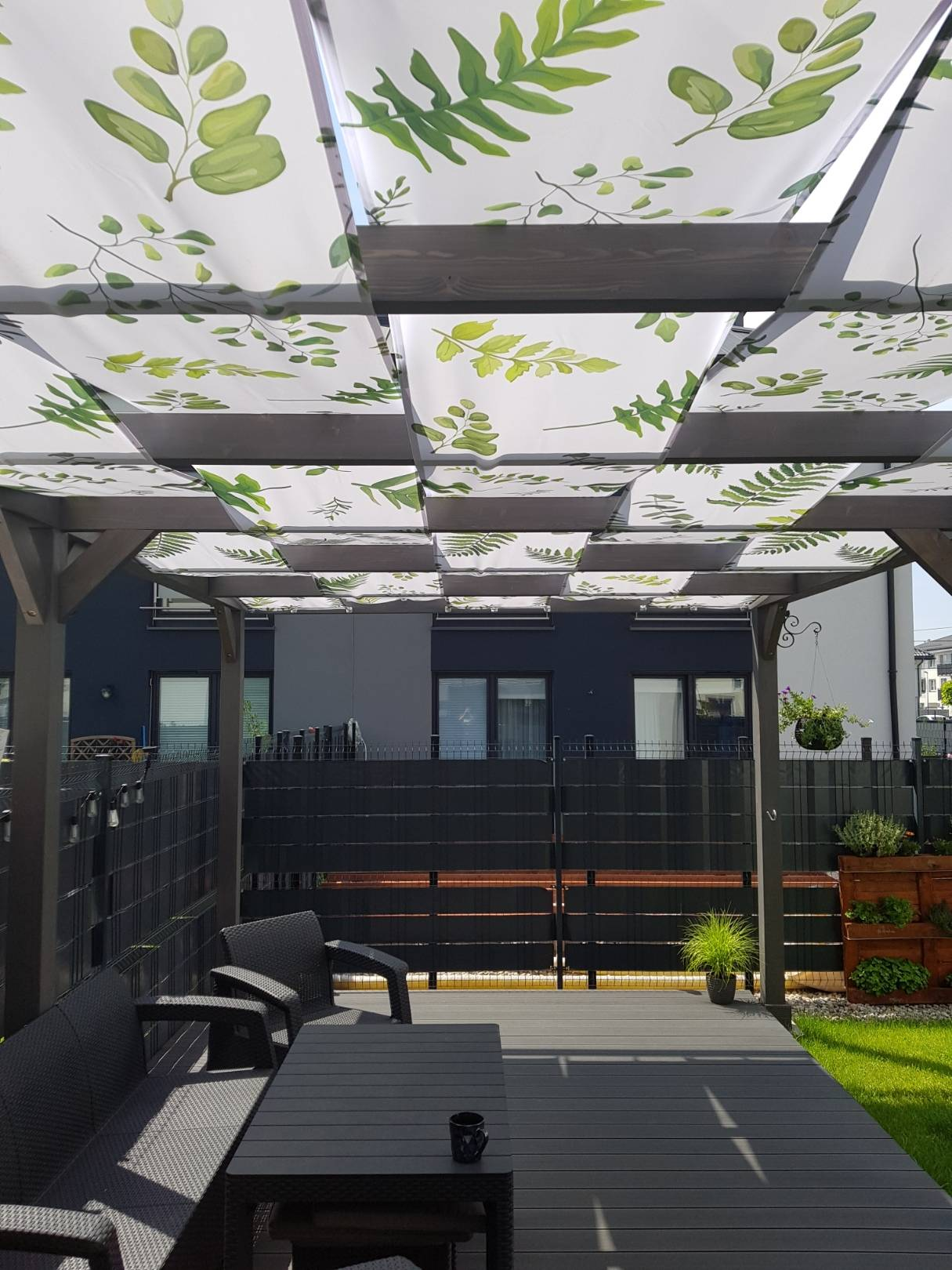 A backyard patio featuring a modern dark-colored deck and furniture under a pergola with a decorative white canopy featuring green leaf prints.