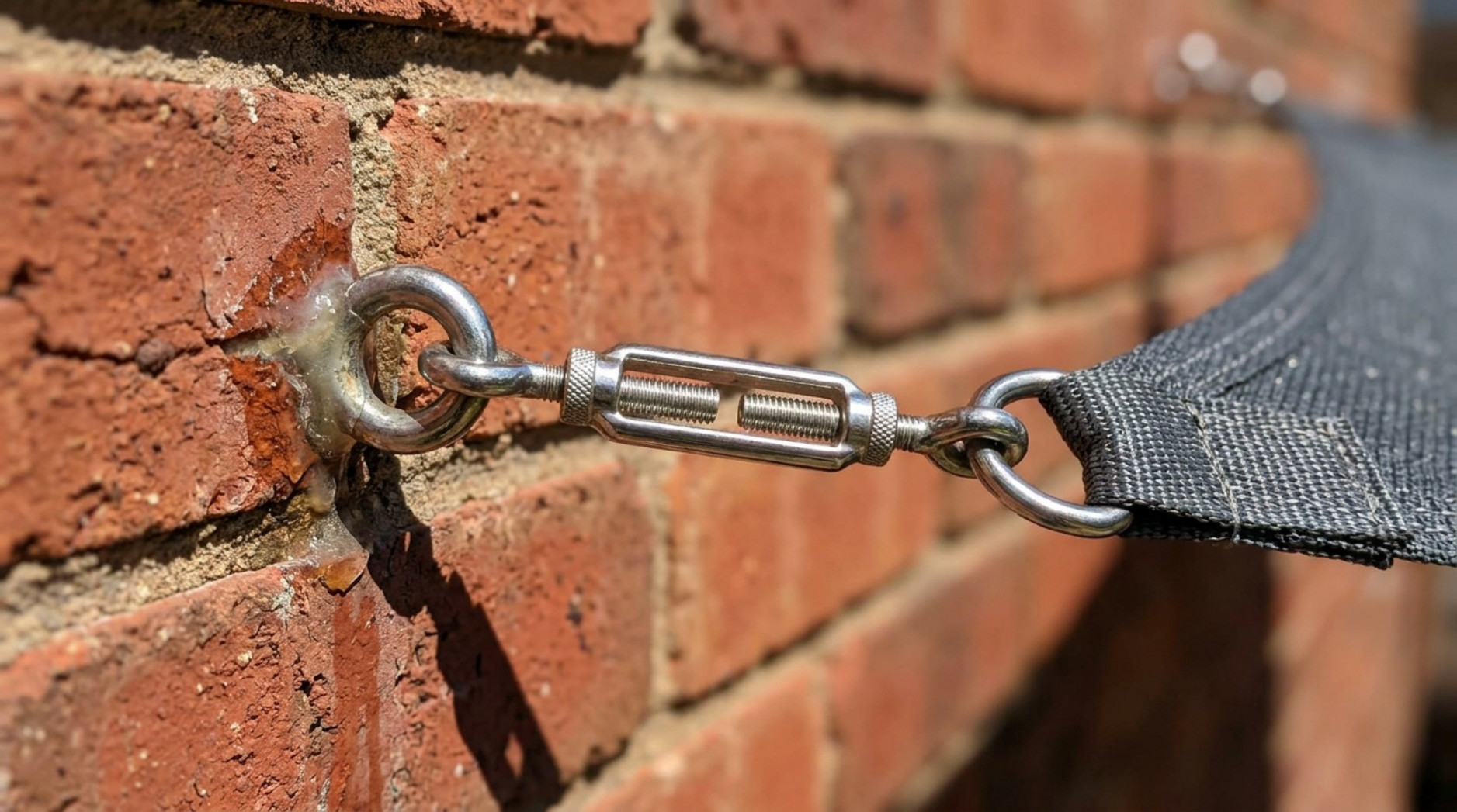 Metal turnbuckle securing a dark shade sail to an eye bolt fixed into a brick wall.