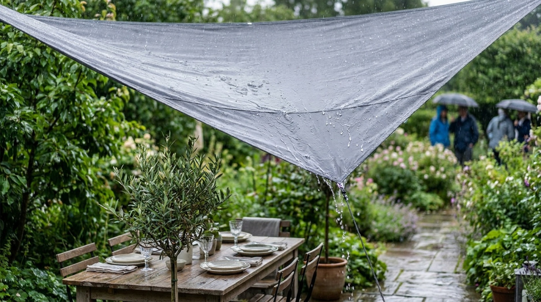 A grey waterproof sun sail stretched tight over an outdoor wooden dining table while rain pours down in a lush green garden.